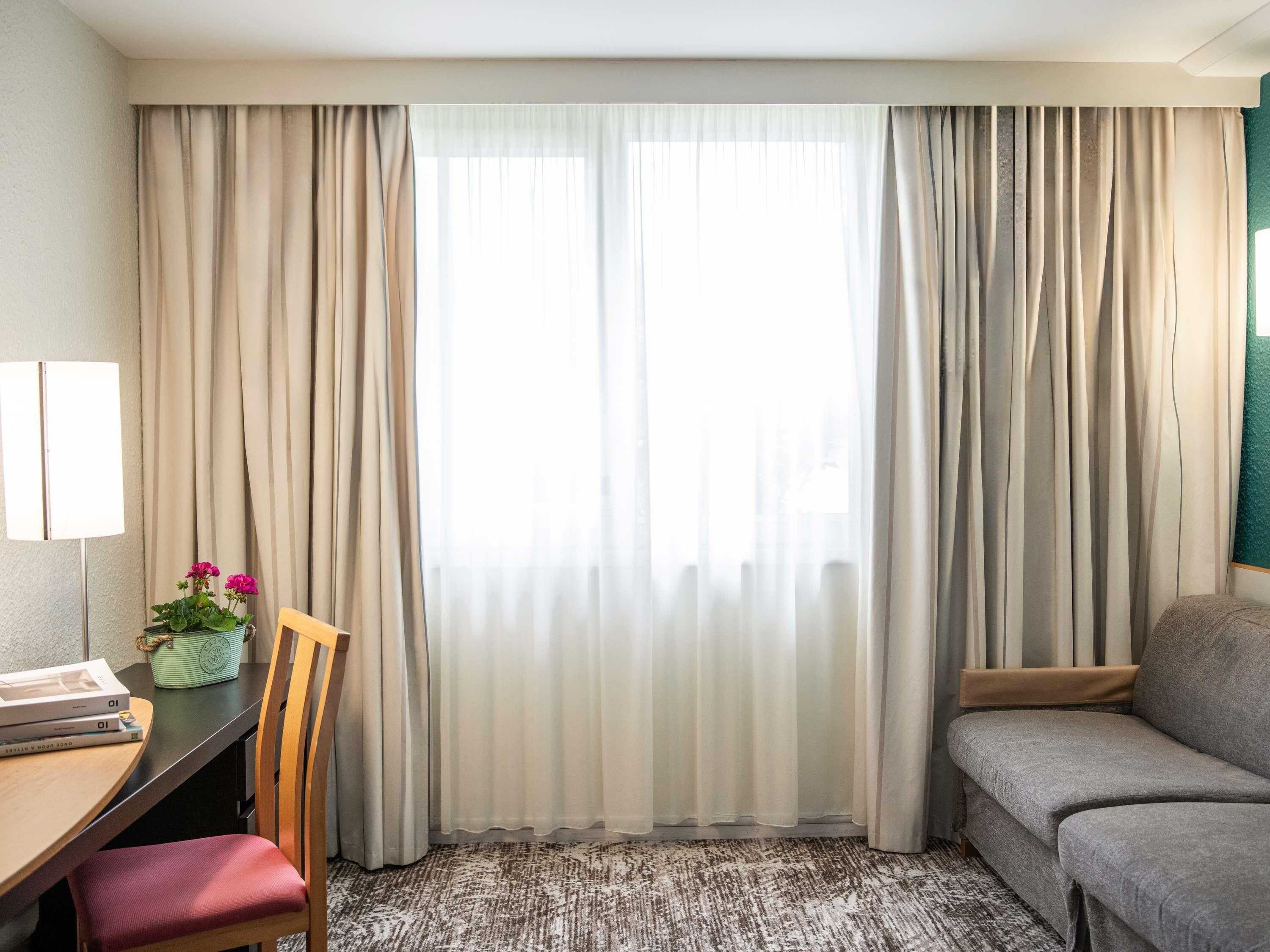 Ein Hotelzimmer mit Schreibtisch, Stuhl, Sofa und einem großen Fenster mit Vorhängen.