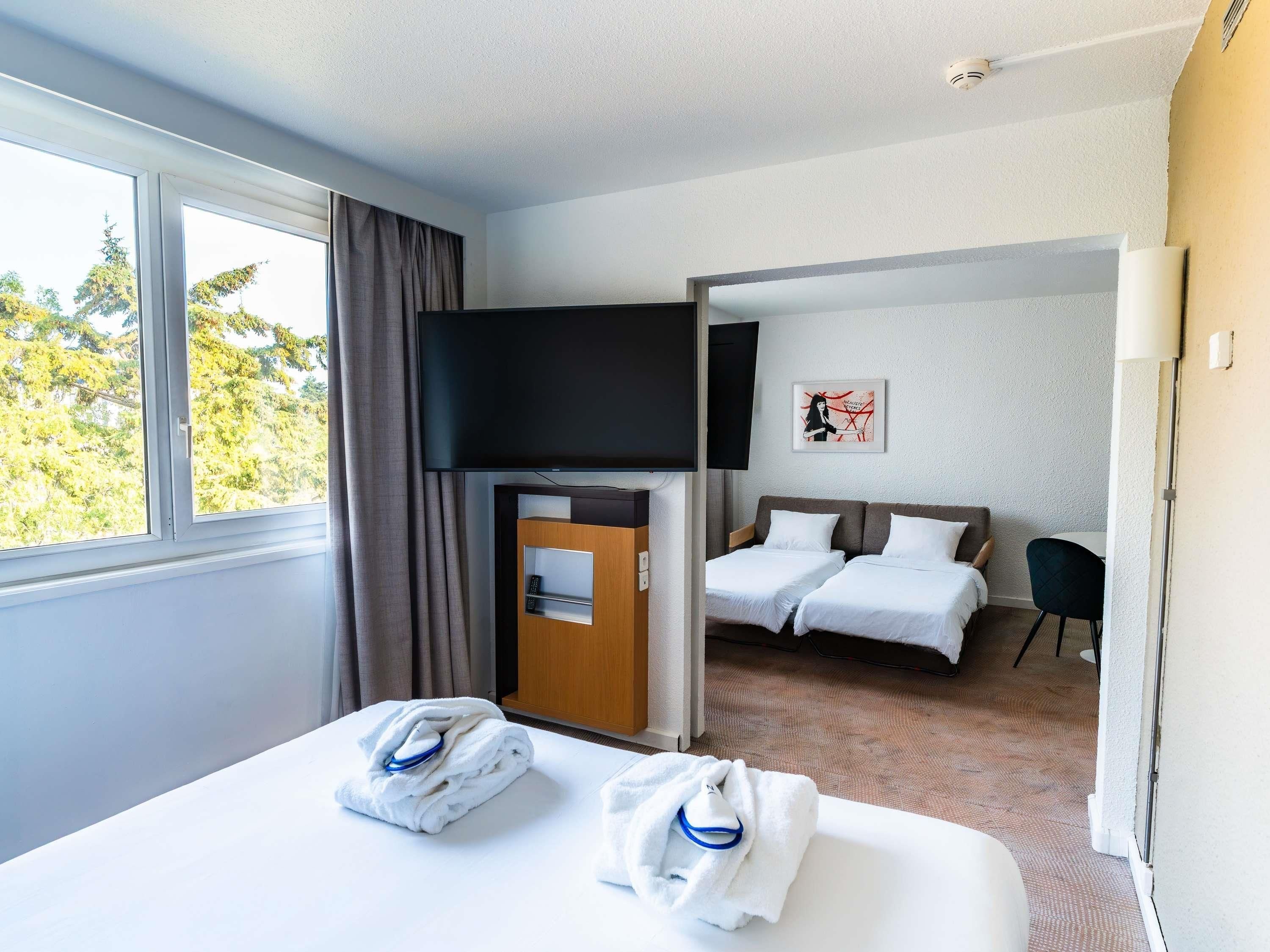 A hotel room with a bed, a TV, a door leading to a balcony, and a view of trees.