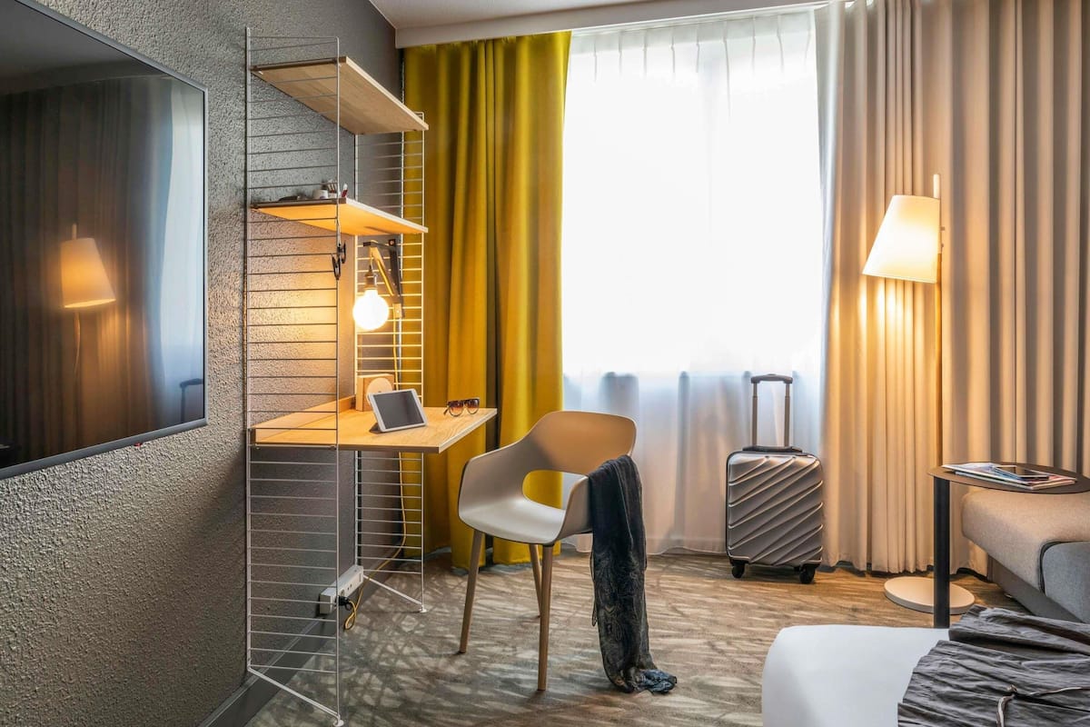A hotel room with a bed, a desk with a laptop, a chair, a suitcase, and a lamp.