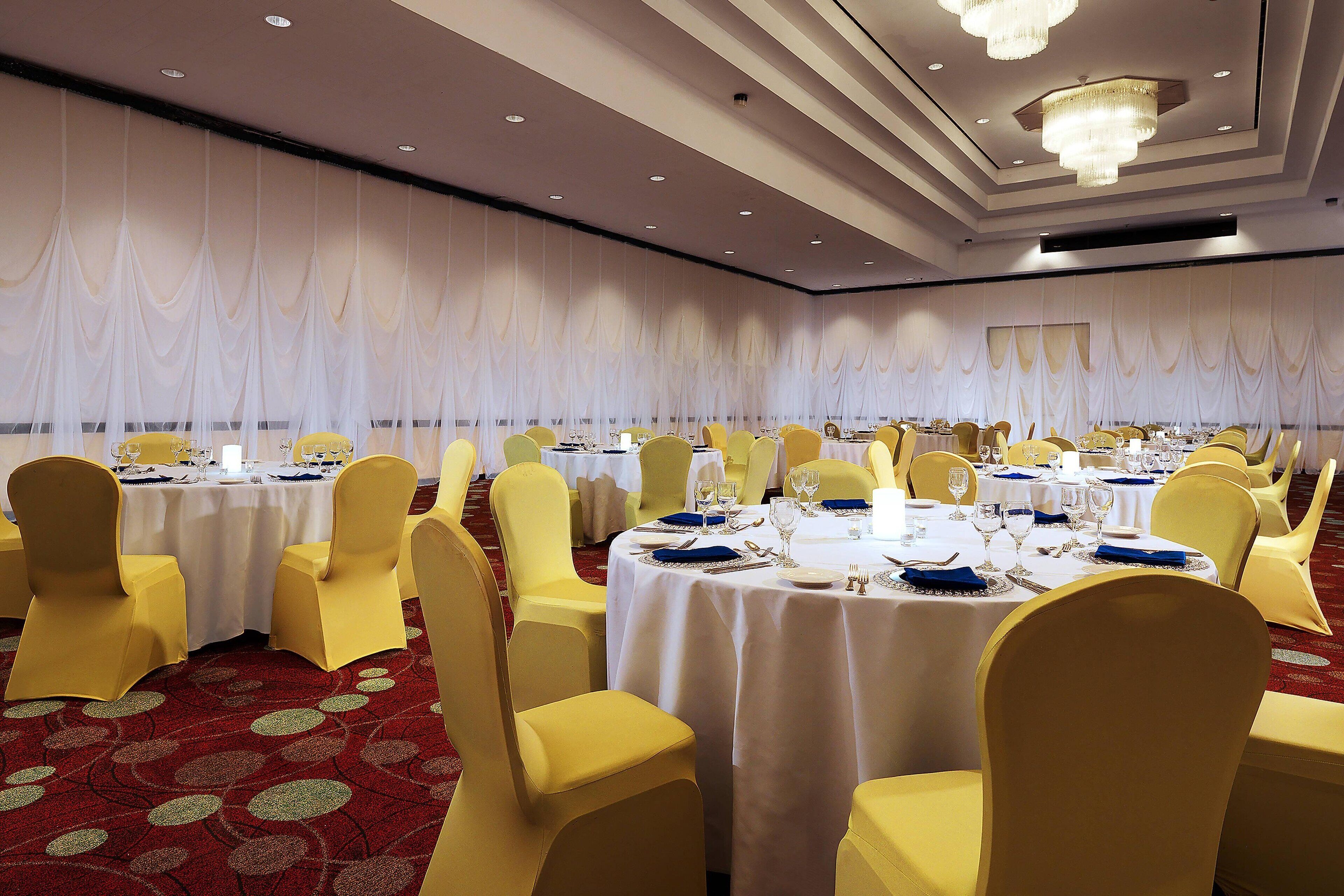 A banquet hall with round tables, yellow chairs, and white tablecloths.