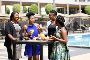 A group of people around a table with drinks and food, near a pool area with umbrellas and lounge chairs.