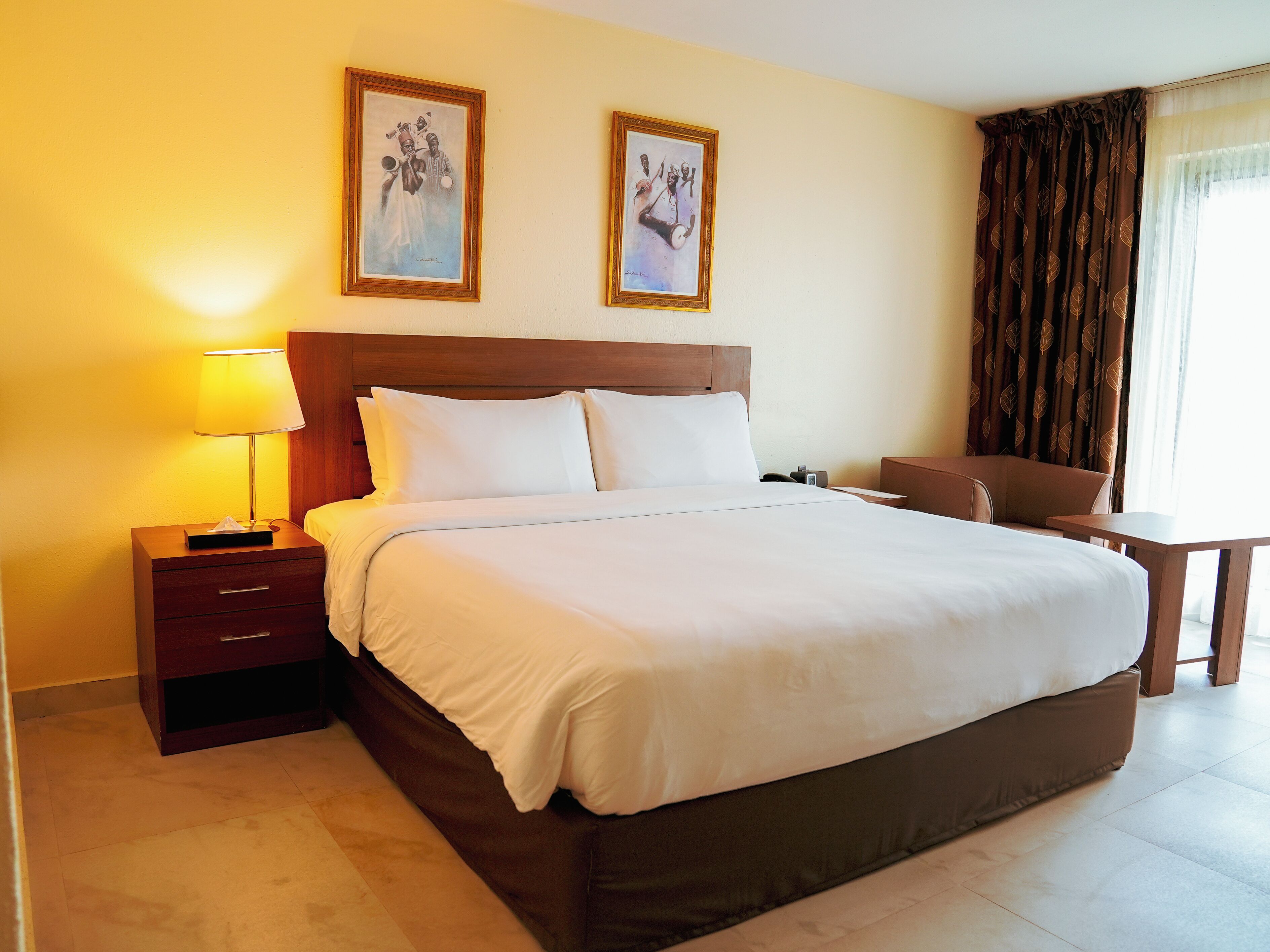 A hotel room with a large bed, a bedside table with a lamp, a chair, a small table, and two framed pictures on the wall.