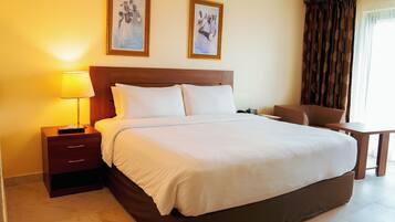 A hotel room with a large bed, a bedside table with a lamp, a chair, a small table, and two framed pictures on the wall.