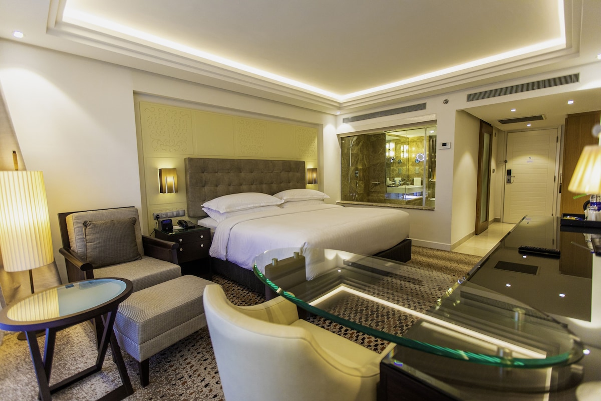 Una habitación de hotel con una cama, un escritorio, una silla, una mesa de centro de vidrio y un espejo.