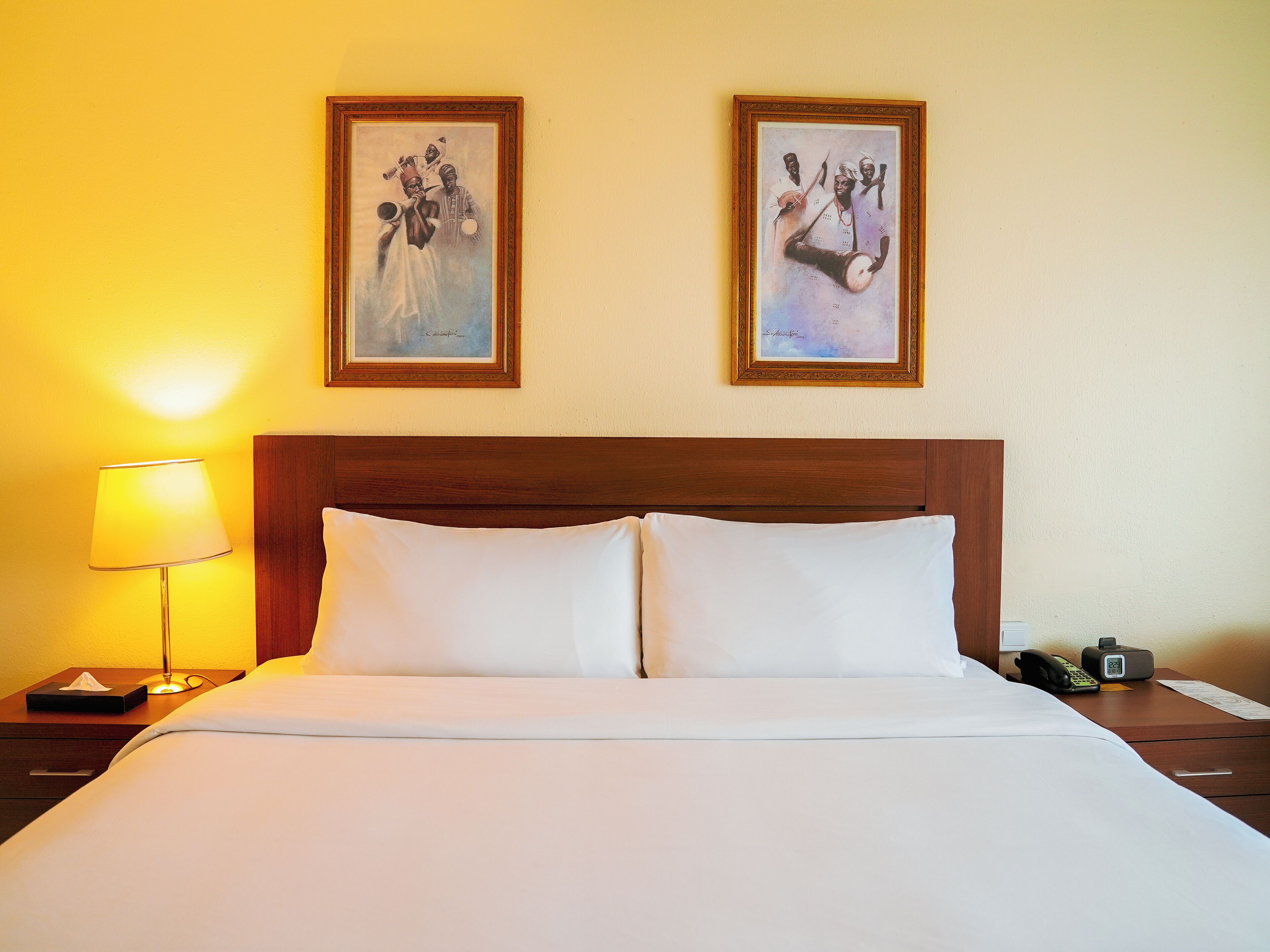 A hotel room with a wooden headboard, two framed artworks on the wall, a bedside lamp, a telephone, and a nightstand with a tissue box.