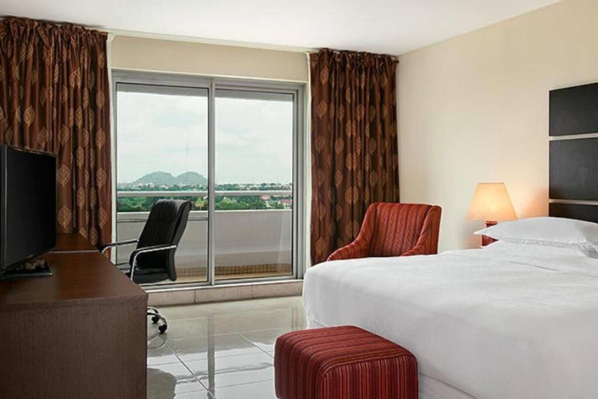 A hotel room with a bed, a TV, a chair, a desk, a lamp, and a view of the outdoors.