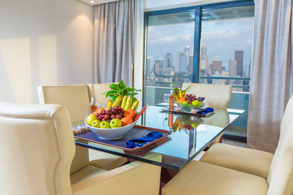 A dining table with a fruit bowl and a tray, two chairs, and a city view through large windows.
