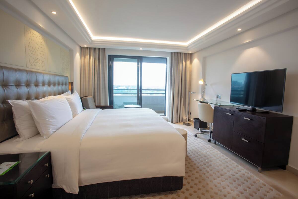 A hotel room with a large bed, a flat-screen TV, a desk with a lamp, and a view of the city through the window.