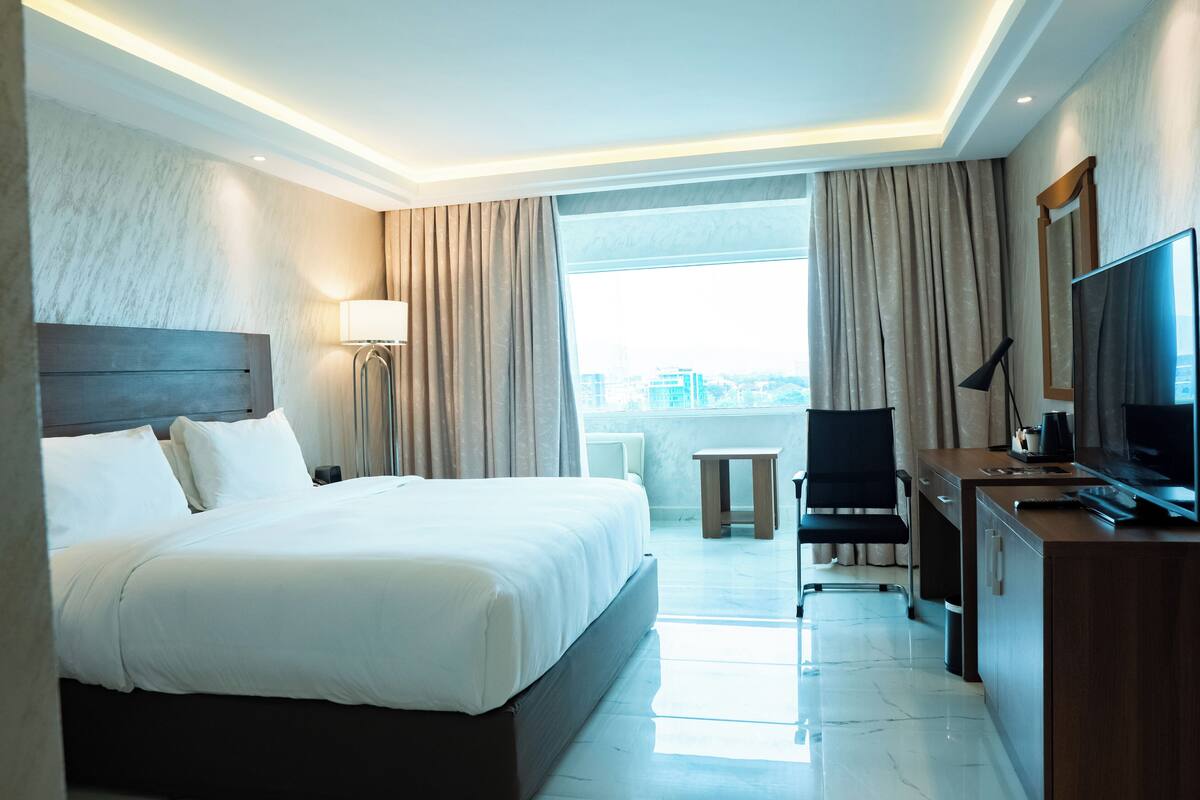 A hotel room with a large bed, a desk with a TV, a chair, and a view of the city through large windows.