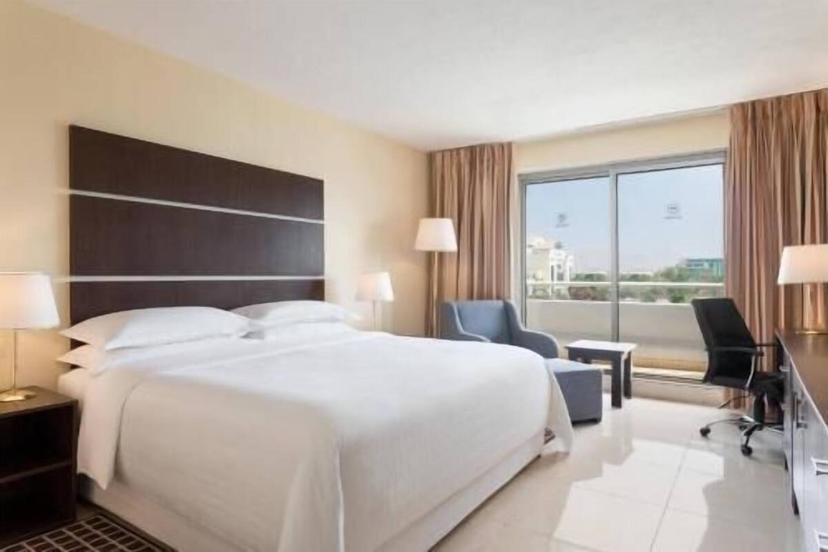 A hotel room with a large bed, a desk, a chair, and a view of the city through the window.