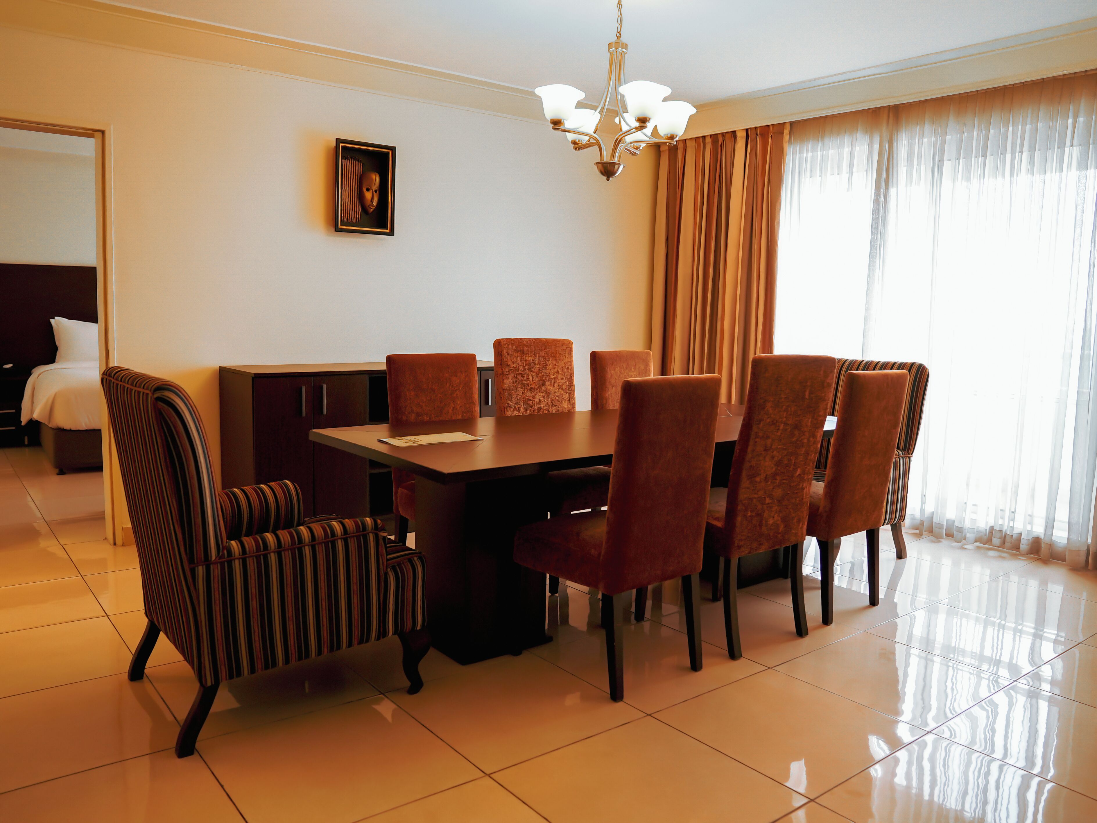 A hotel dining area with a rectangular table, six chairs, a chandelier, and a large window with curtains.