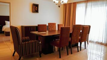 A hotel dining area with a rectangular table, six chairs, a chandelier, and a large window with curtains.