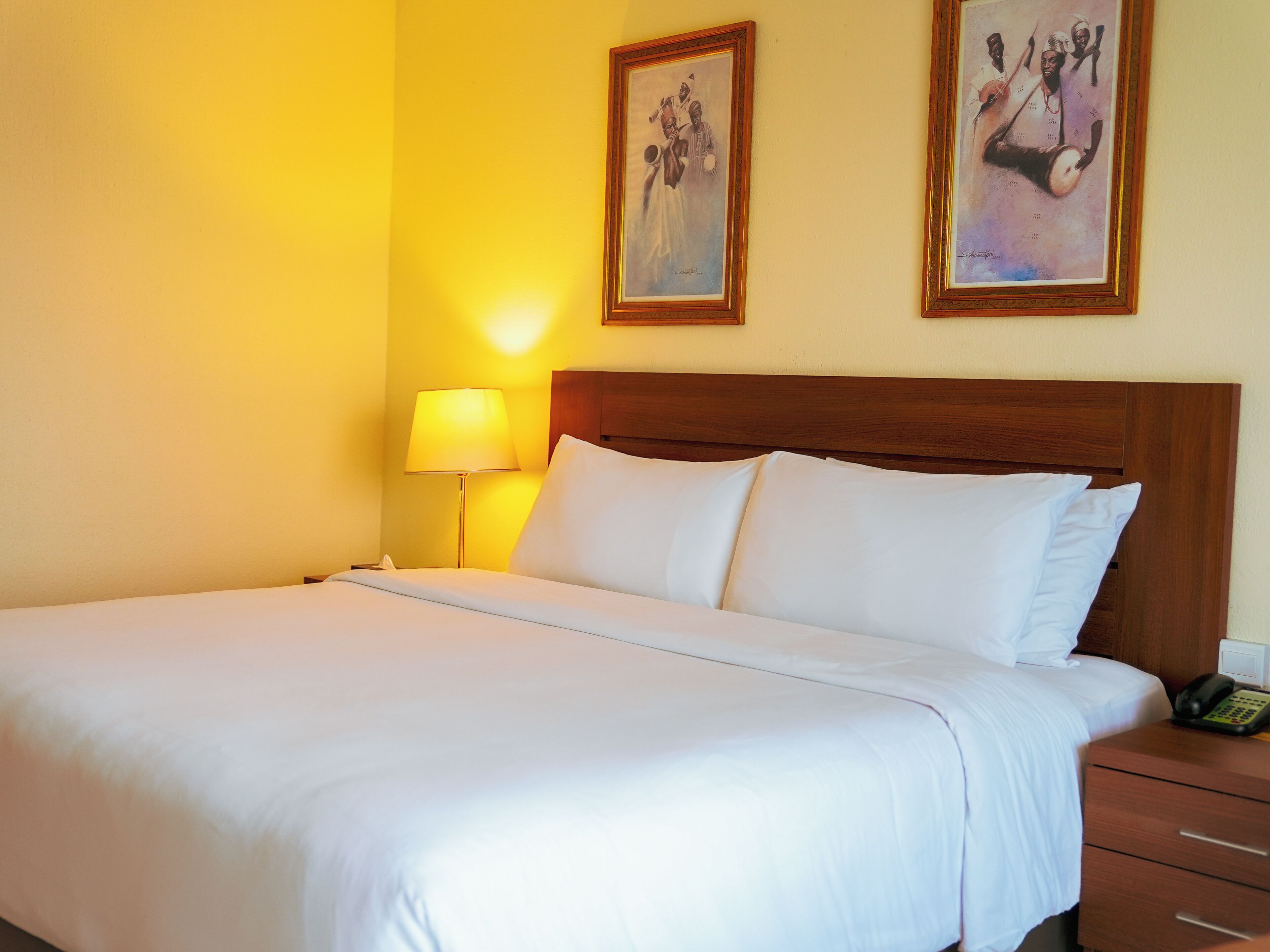 Camera d'hotel con un letto grande, una testiera in legno, una lampada da comodino e due opere d'arte appese alla parete.