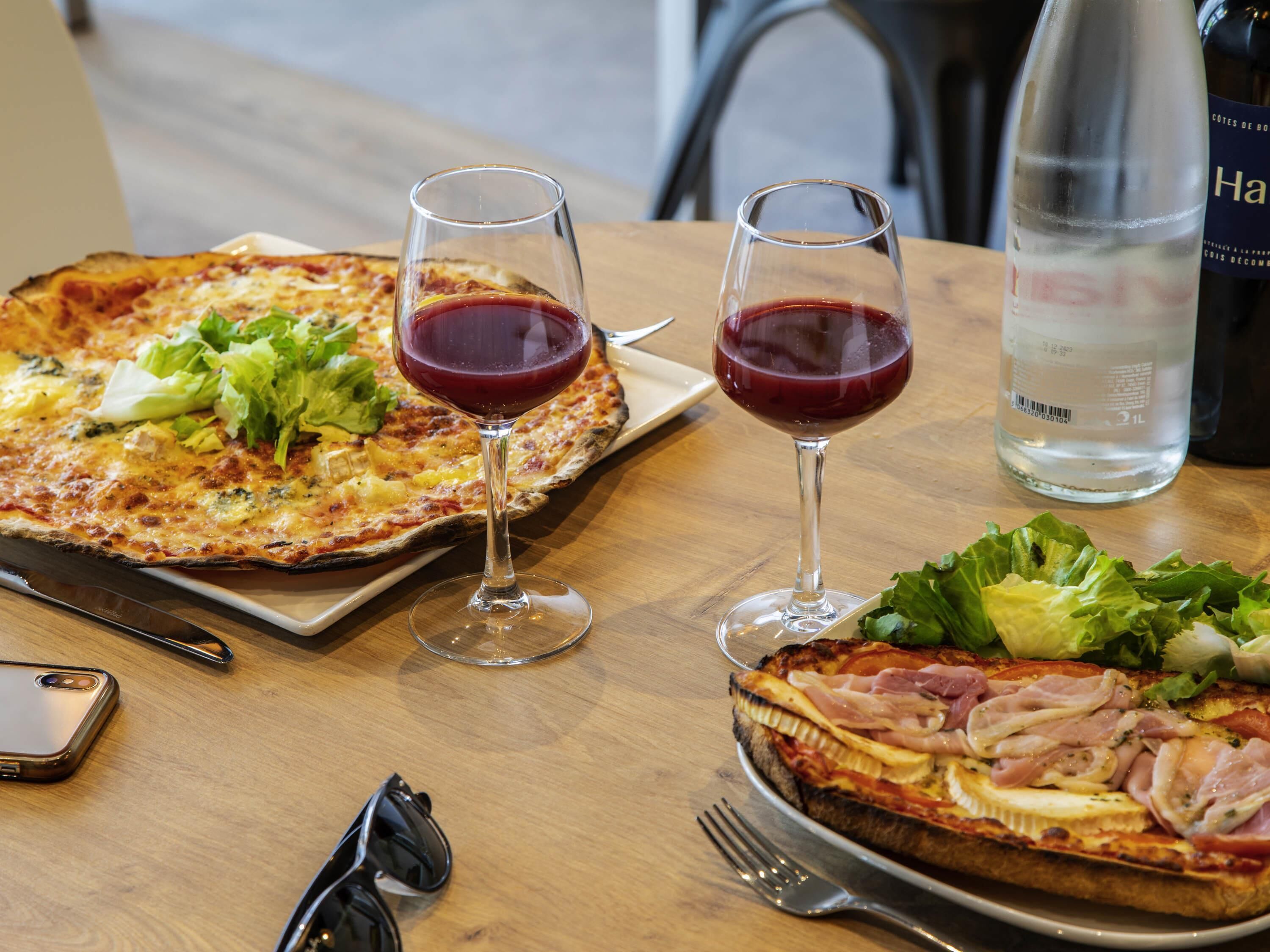 A wooden table with a pizza, two glasses of red wine, a bottle of water, a smartphone, and a pair of sunglasses.