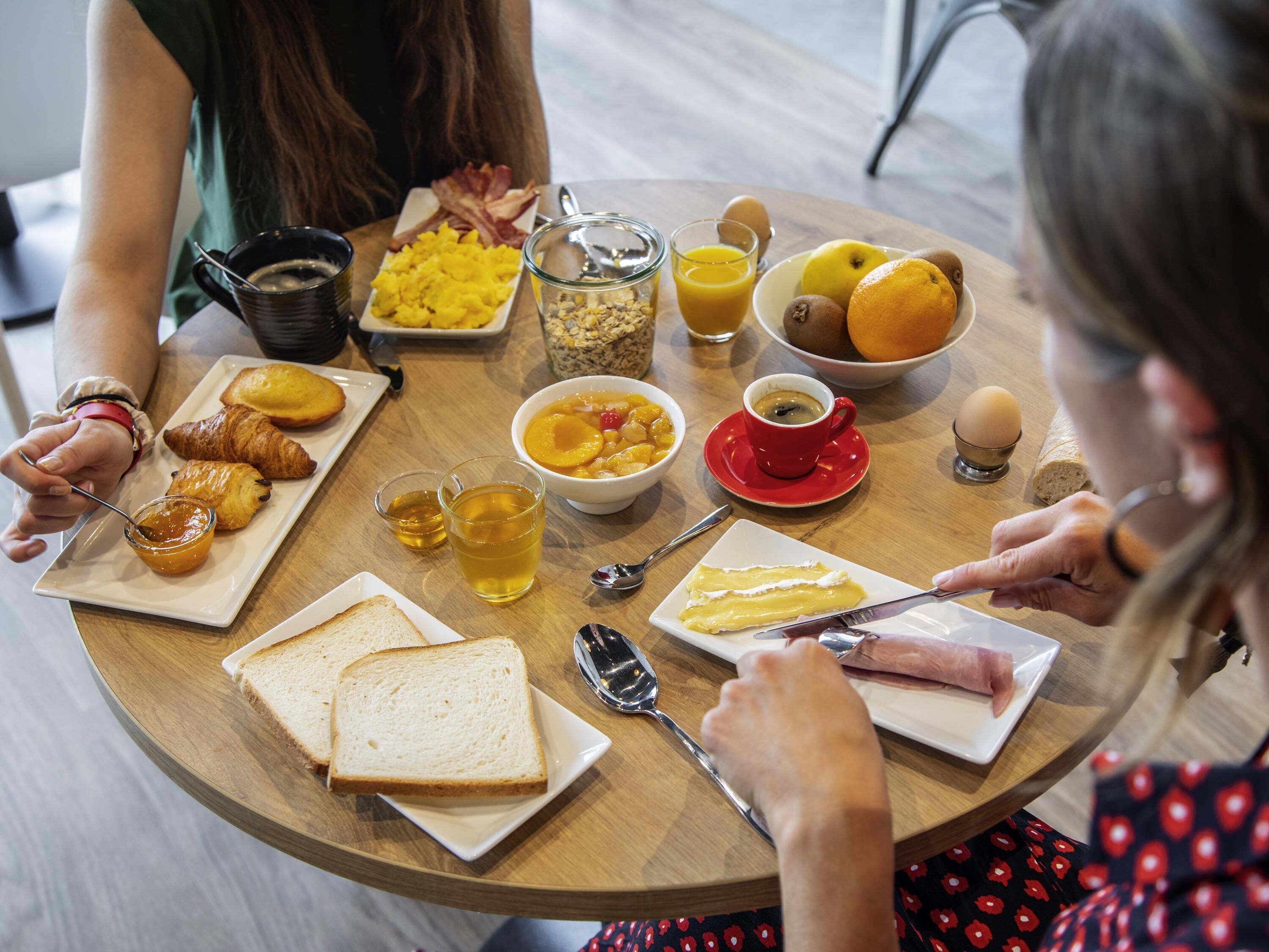 A round wooden table with breakfast items including croissants, toast, a bowl of fruit, a cup of coffee, and a glass of juice.