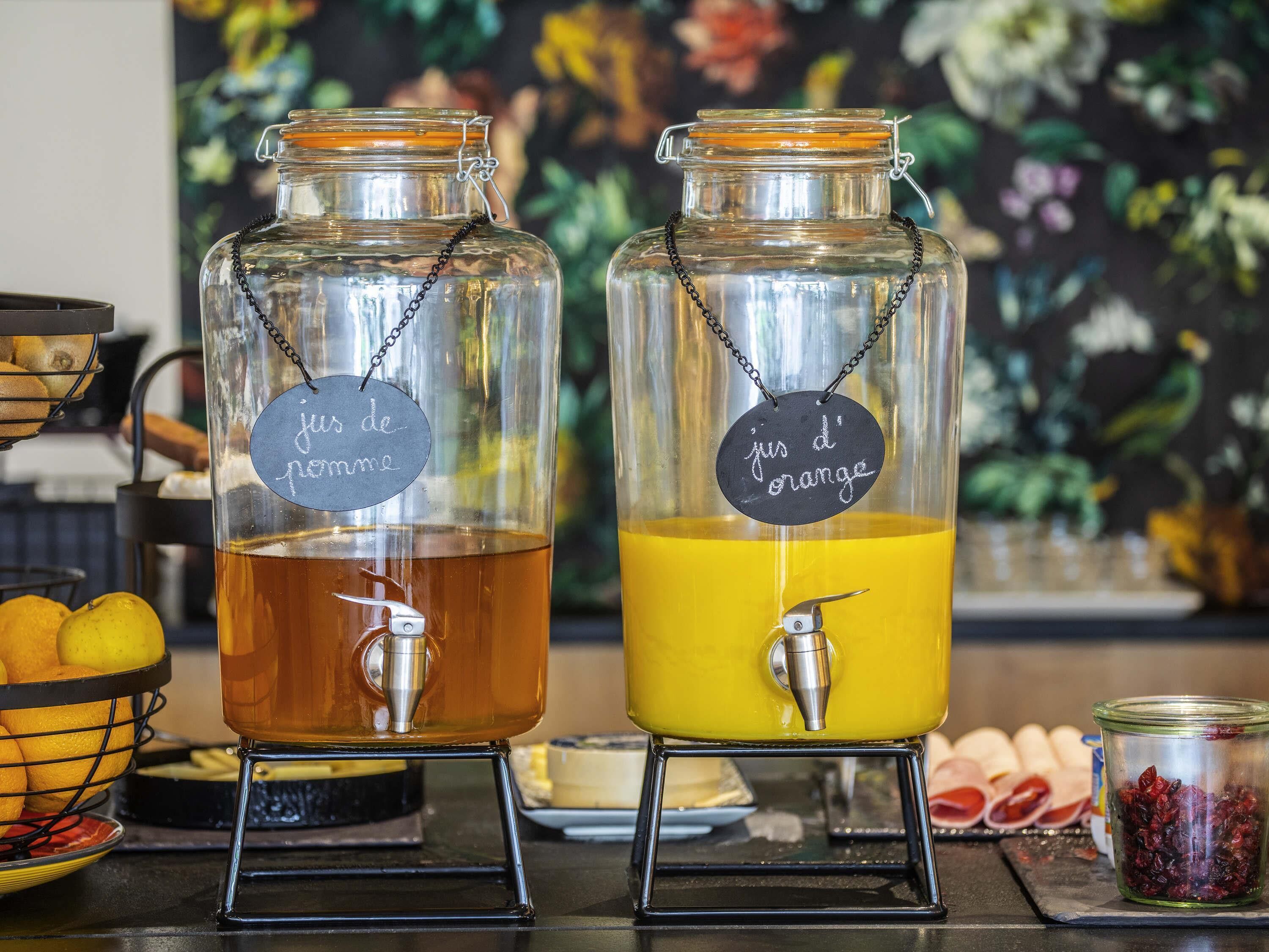 Two glass dispensers with metal chains and labels, one labeled 'jus de pomme' and the other 'jus d'orange,' placed on a counter with fruits and a floral backdrop.