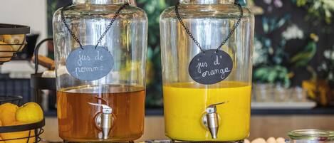 Two glass dispensers with metal chains and labels, one labeled 'jus de pomme' and the other 'jus d'orange,' placed on a counter with fruits and a floral backdrop.