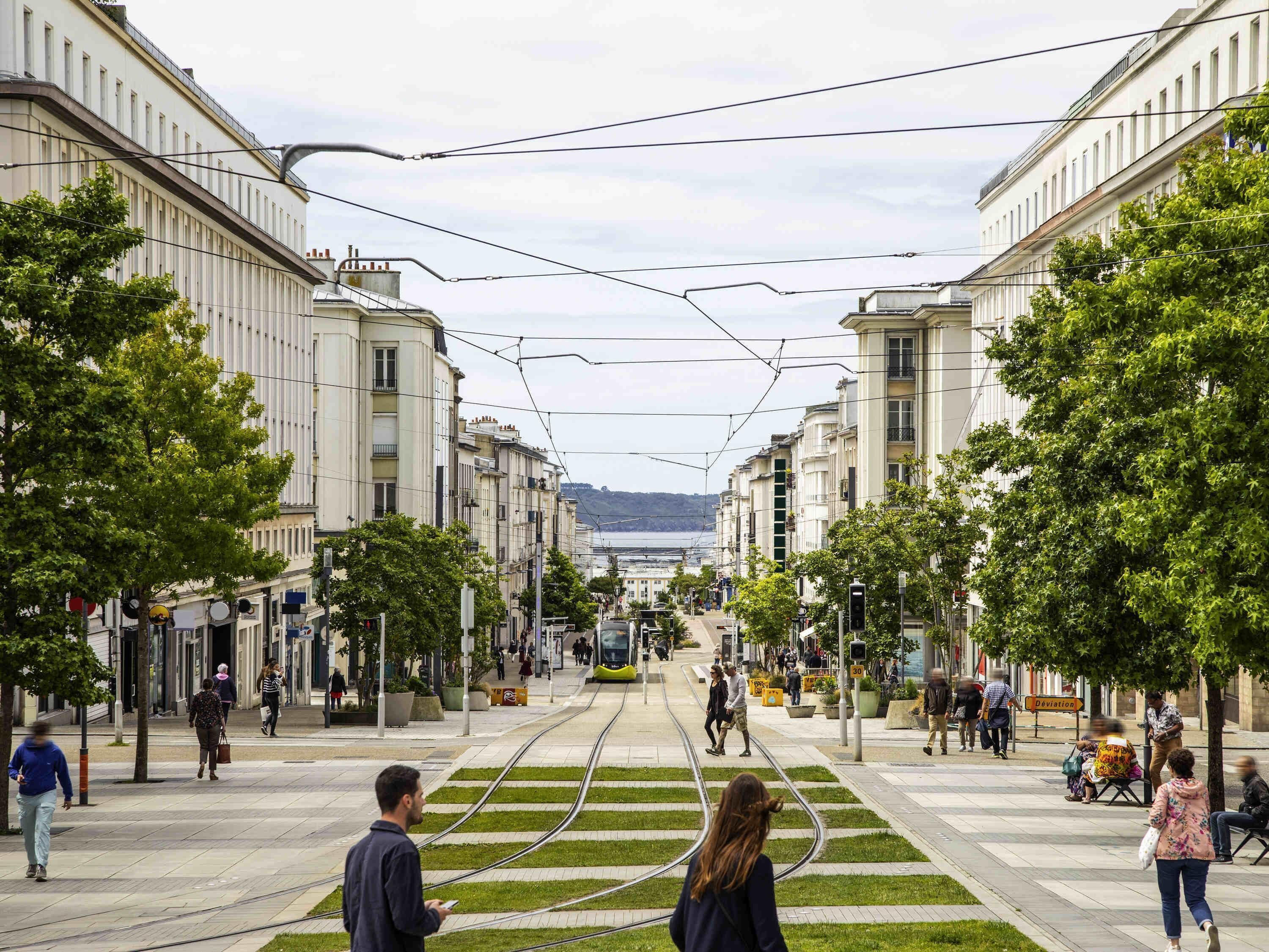 A tram line with overhead wires, a pedestrian walkway, and buildings on either side.