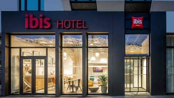 Ein Hotel Eingang mit Glastüren, einem Schild mit der Aufschrift 'ibis HOTEL' und einem modernen Innenraum, der durch die Türen sichtbar ist.