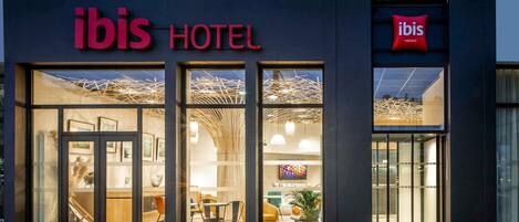 La entrada de un hotel con puertas de vidrio, un cartel que dice 'ibis HOTEL' y un interior moderno visible a través de las puertas.