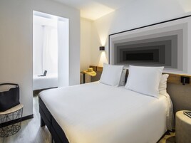 A hotel room with a large bed, a desk, a chair, a window, and a wall-mounted TV.