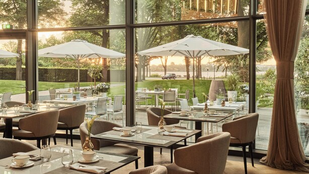 A restaurant with outdoor seating, large windows, and a view of a garden and lake. - The Fontenay (Hamburg)