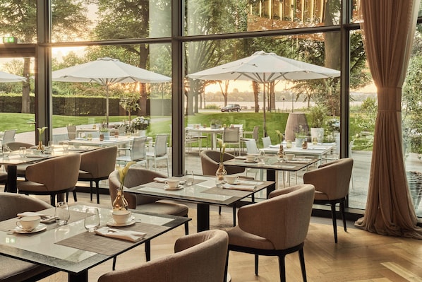 A restaurant with outdoor seating, large windows, and a view of a garden and lake. - The Fontenay (Hamburg)