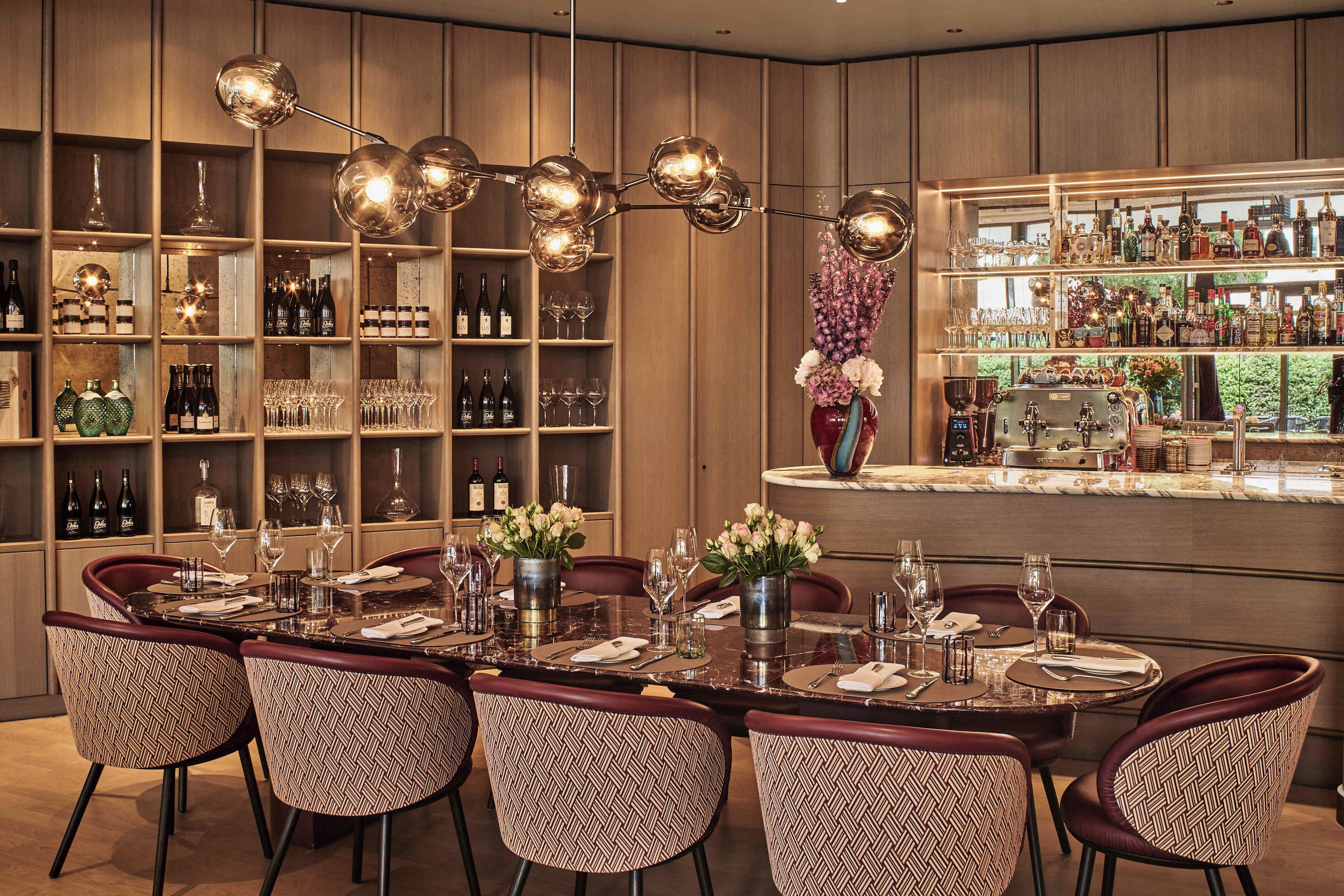 A dining area with a long table set for six, surrounded by patterned chairs. There are shelves with bottles and glasses, and a bar area with a marble countertop.