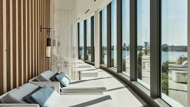 A modern lounge area with white sofas and cushions, large windows offering a view of a river and cityscape. - The Fontenay (Hamburg)