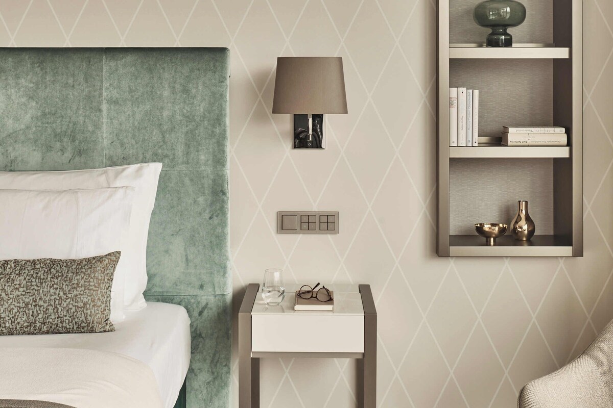 A modern bedroom with a green upholstered headboard, a bedside table with a lamp, a bookshelf, and a chair.