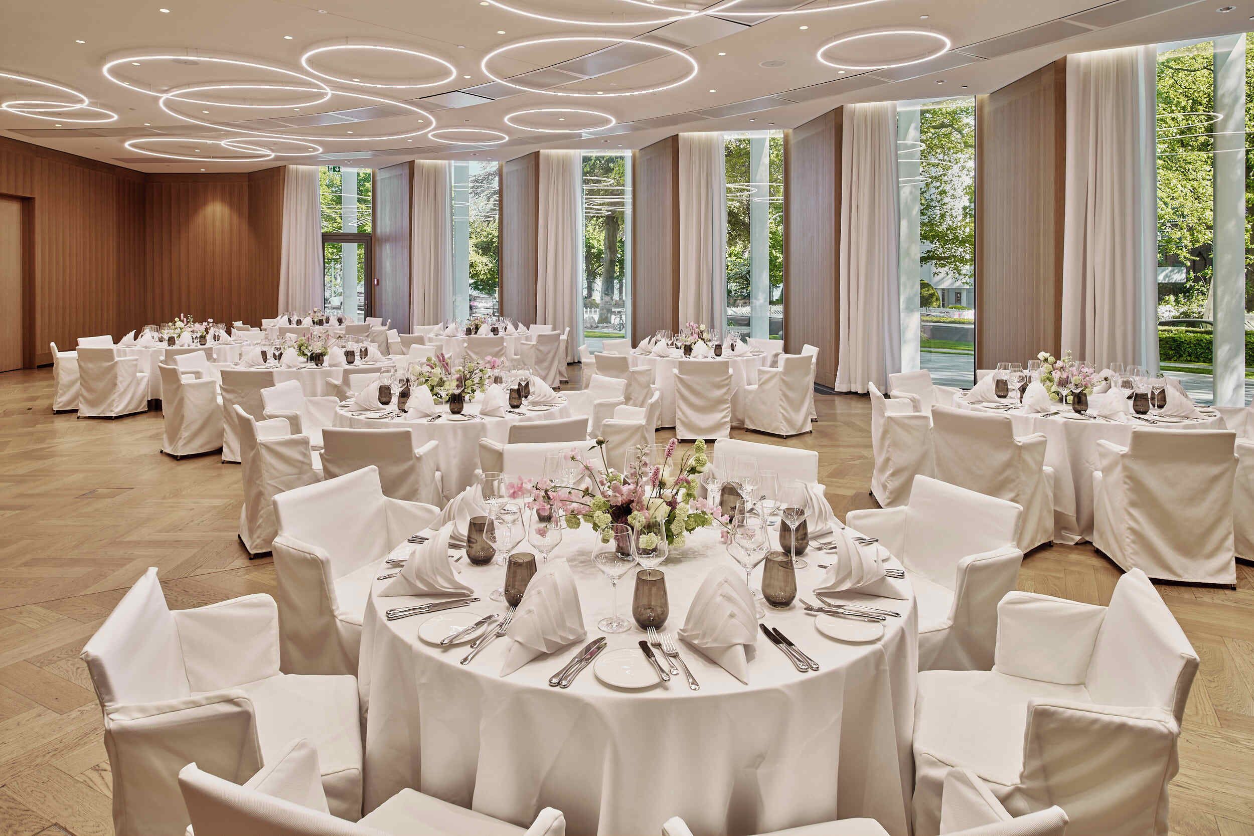 Un salón de banquetes con mesas redondas dispuestas para comer, ventanas amplias y un diseño moderno del techo.