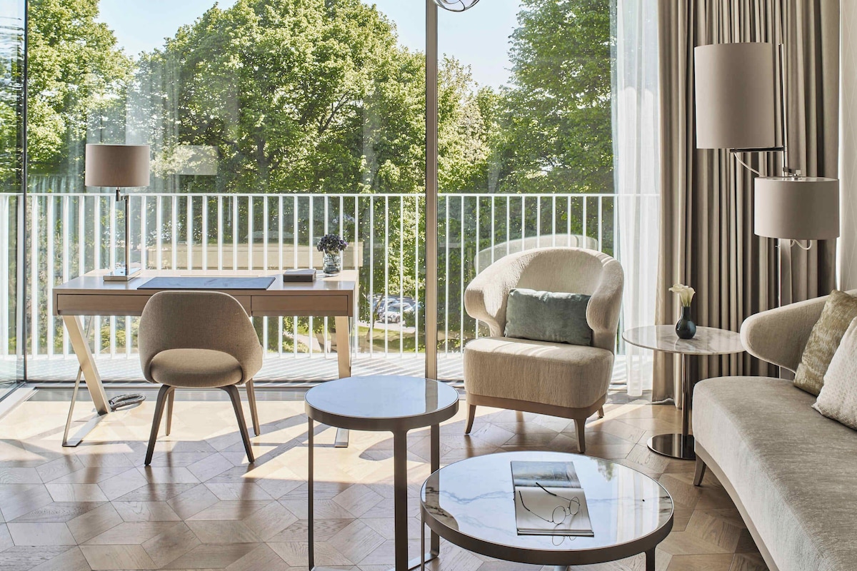 Una sala de estar moderna con un sofá, sillones, una mesa de centro y un escritorio con una computadora portátil. Se ve un balcón con barandilla y vistas a árboles.