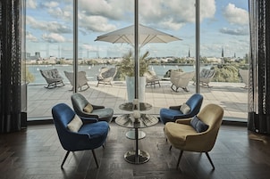 Ein modernes Zimmer mit großen Fenstern, Blick auf eine Stadtlandschaft und einer Terrasse mit Korbmöbeln und einem Tisch.
