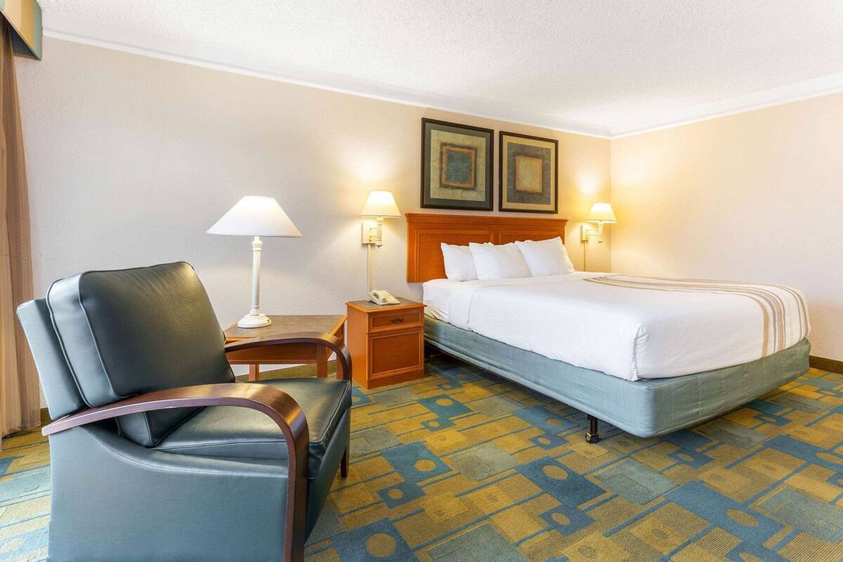 A hotel room with a bed, a chair, a nightstand, a lamp, and two framed pictures on the wall.