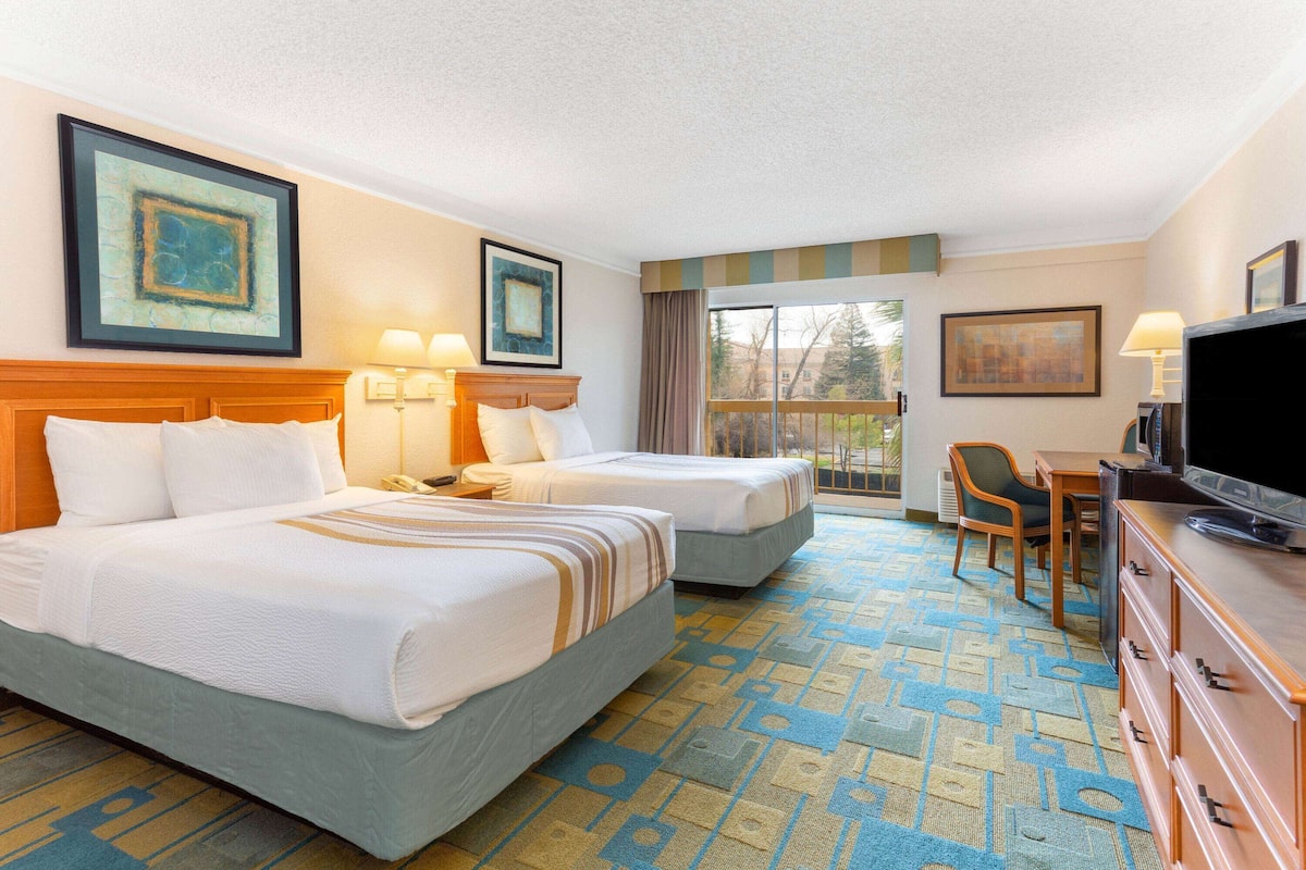 A hotel room with two beds, a desk, a television, and a balcony with a view.