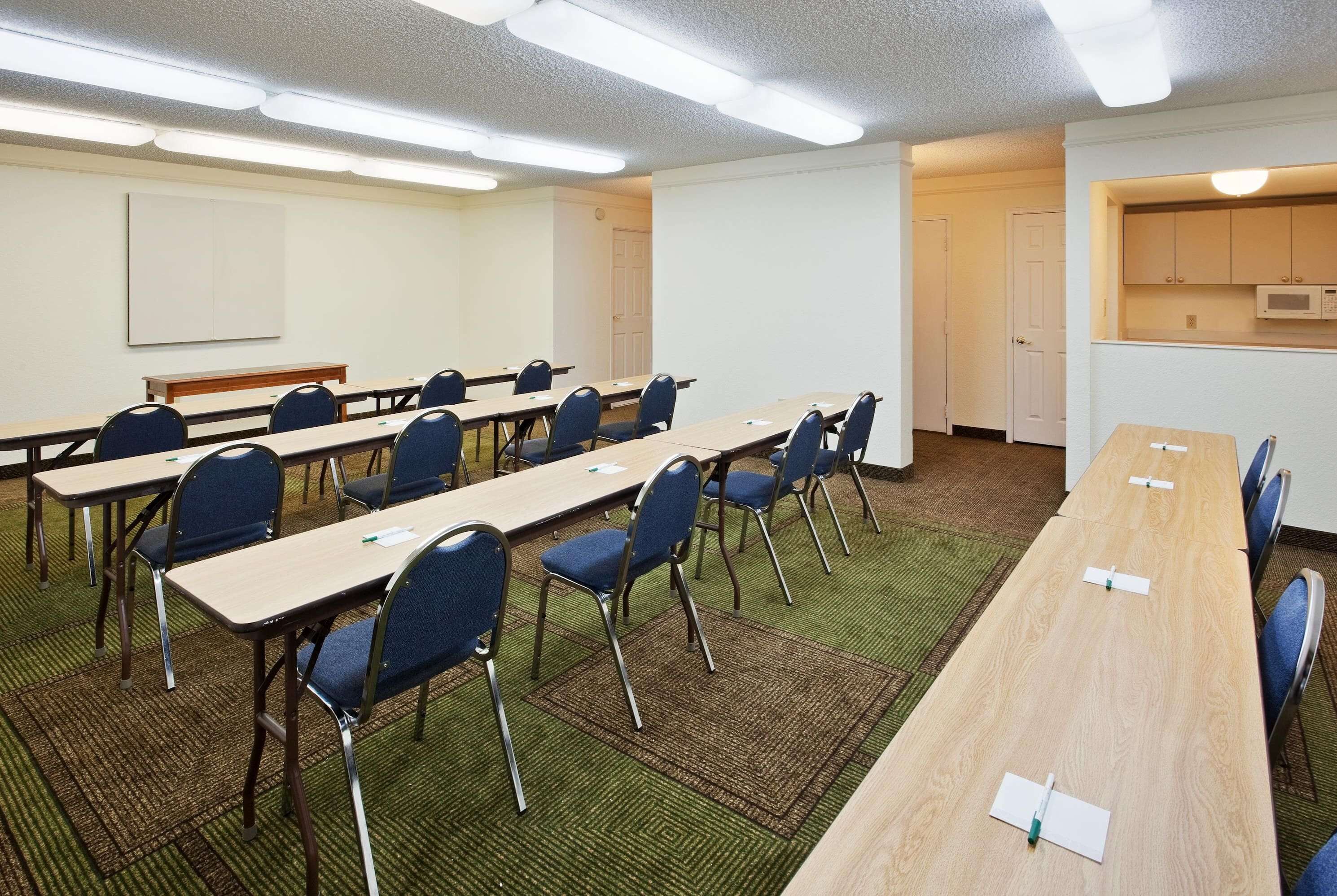 Una sala de conferències amb taules llargues, cadires, una pissarra i una zona de cuina petita.