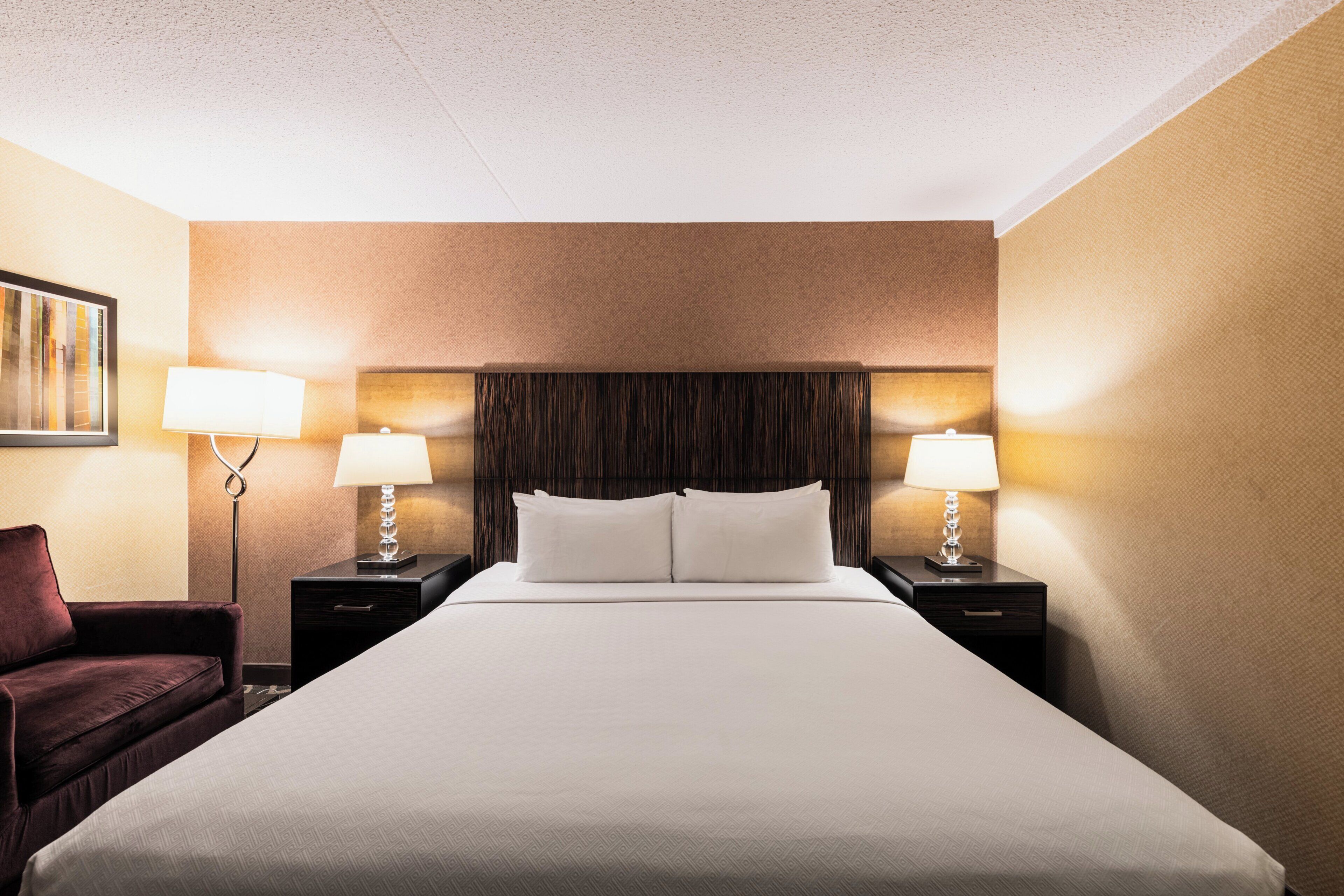 A hotel room with a large bed, two bedside tables with lamps, a dark headboard, and a single chair.