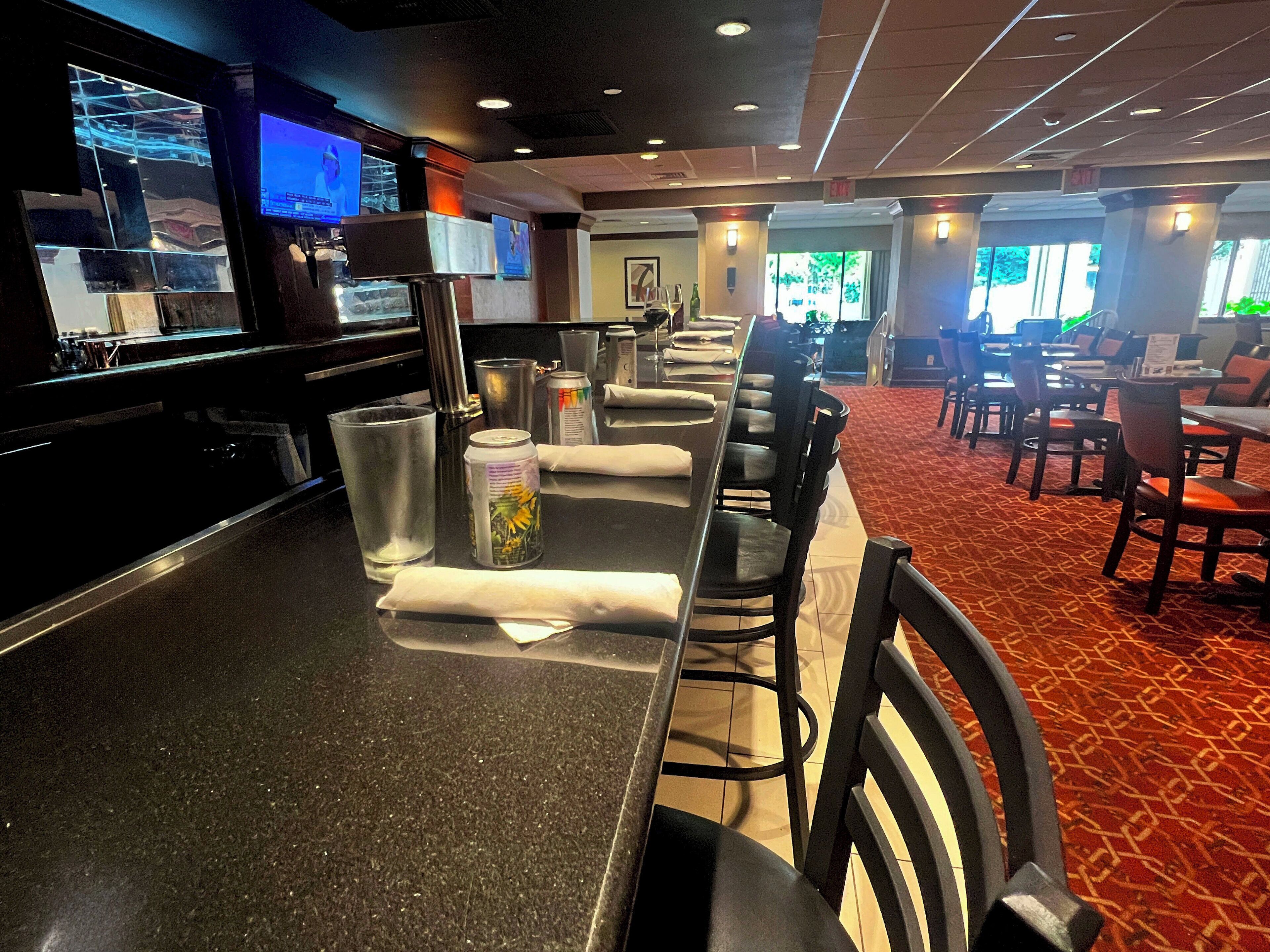 A bar area with a counter, chairs, and tables. There are glasses, a can, and a napkin on the counter. The room has a patterned carpet and a view of the outside.