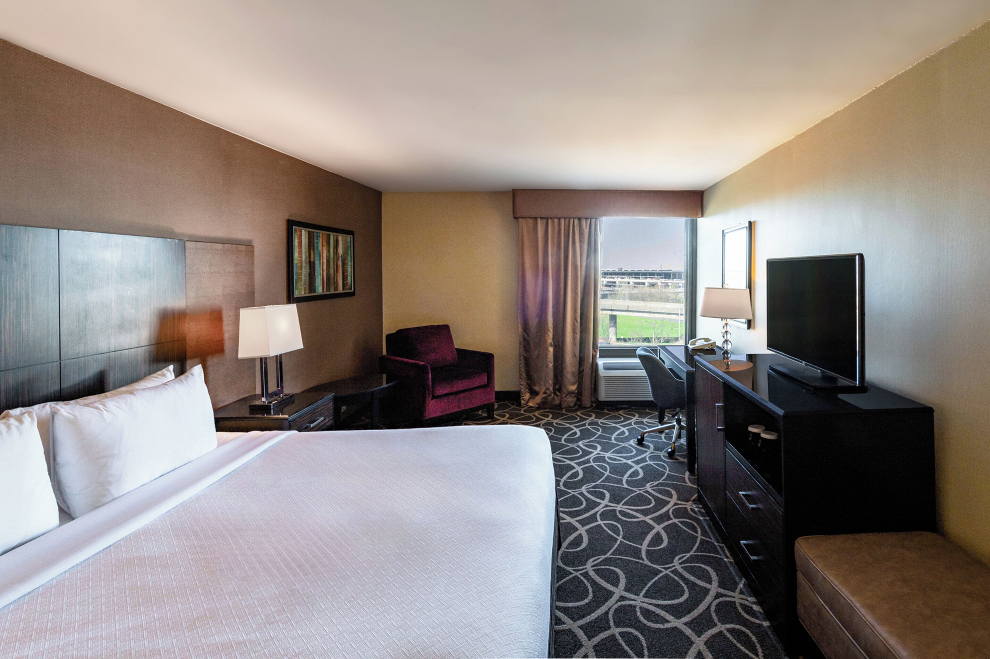 A hotel room with a large bed, a desk, a chair, a TV, and a window with a view.