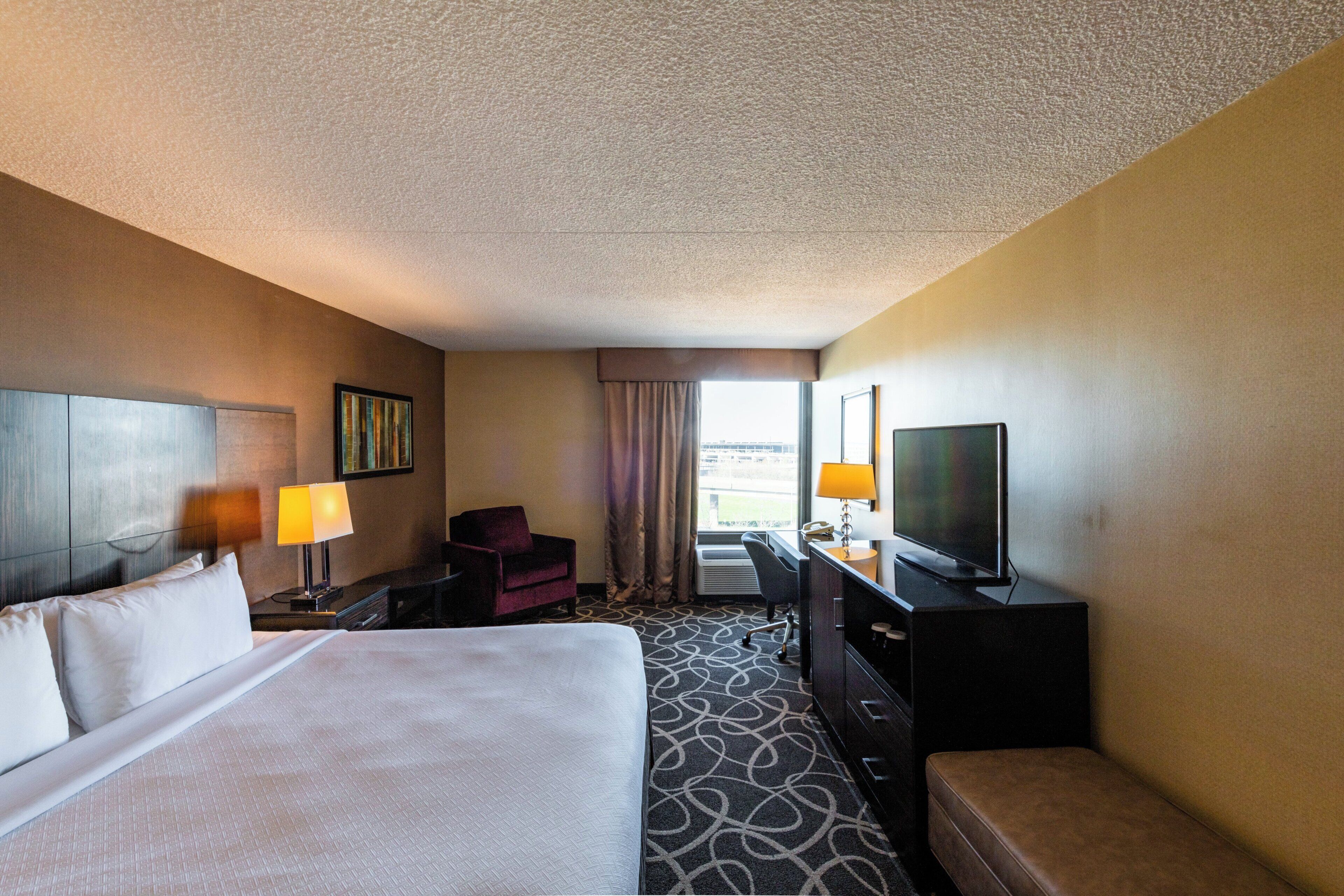 A hotel room with a bed, a desk with a television, a chair, and a window with curtains.