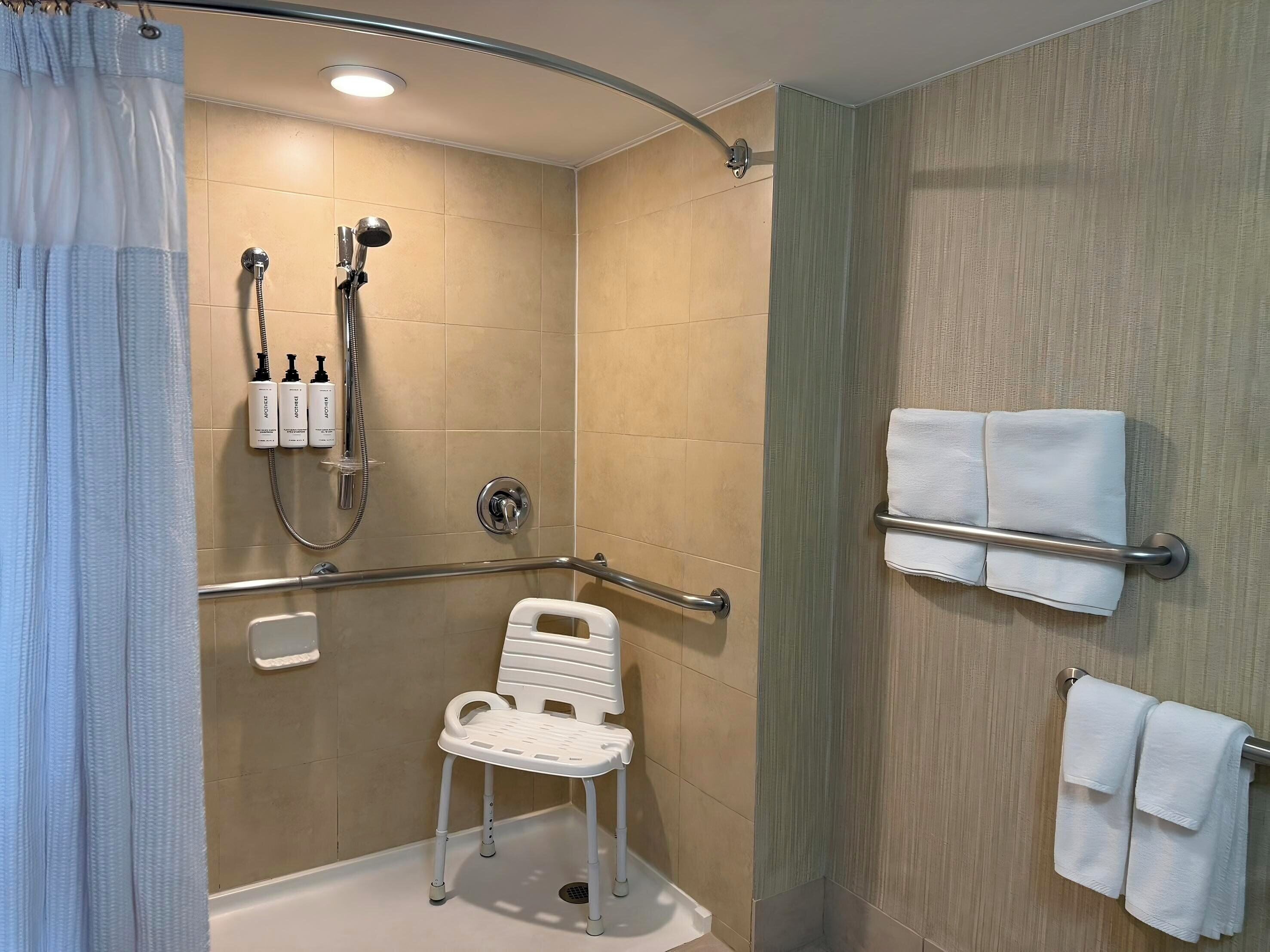 A modern bathroom with a shower, grab bars, a white chair, and towels on a rack.