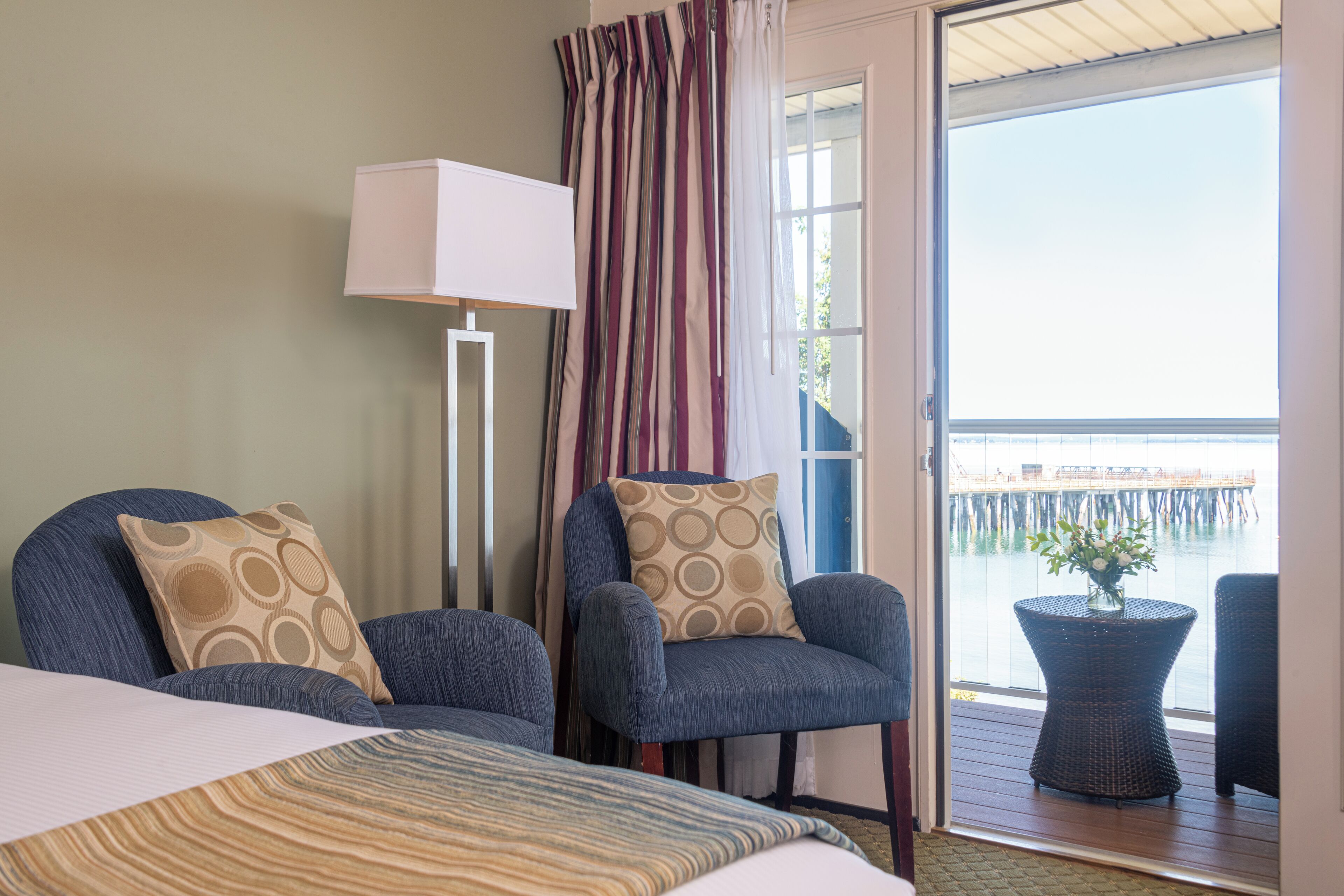 Habitación de hotel con dos sillones, una cama, una lámpara de pie y vistas a un muelle a través de un ventanal grande.