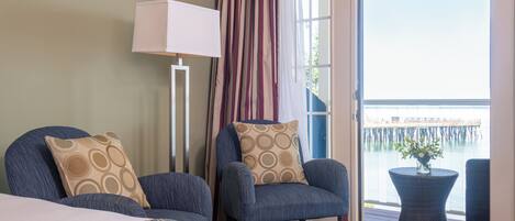A hotel room with two armchairs, a bed, a standing lamp, and a view of a pier through a large window.