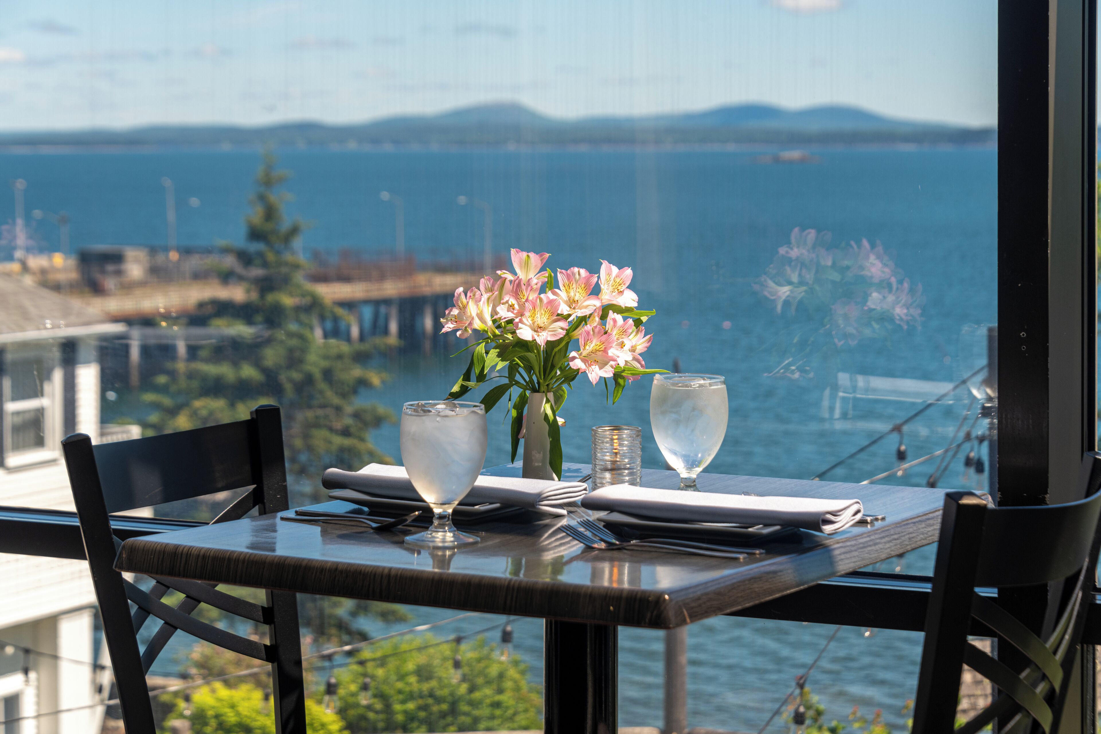 A table set for two with a vase of flowers, two glasses, and cutlery. A view of the sea and a bridge is visible through the window.