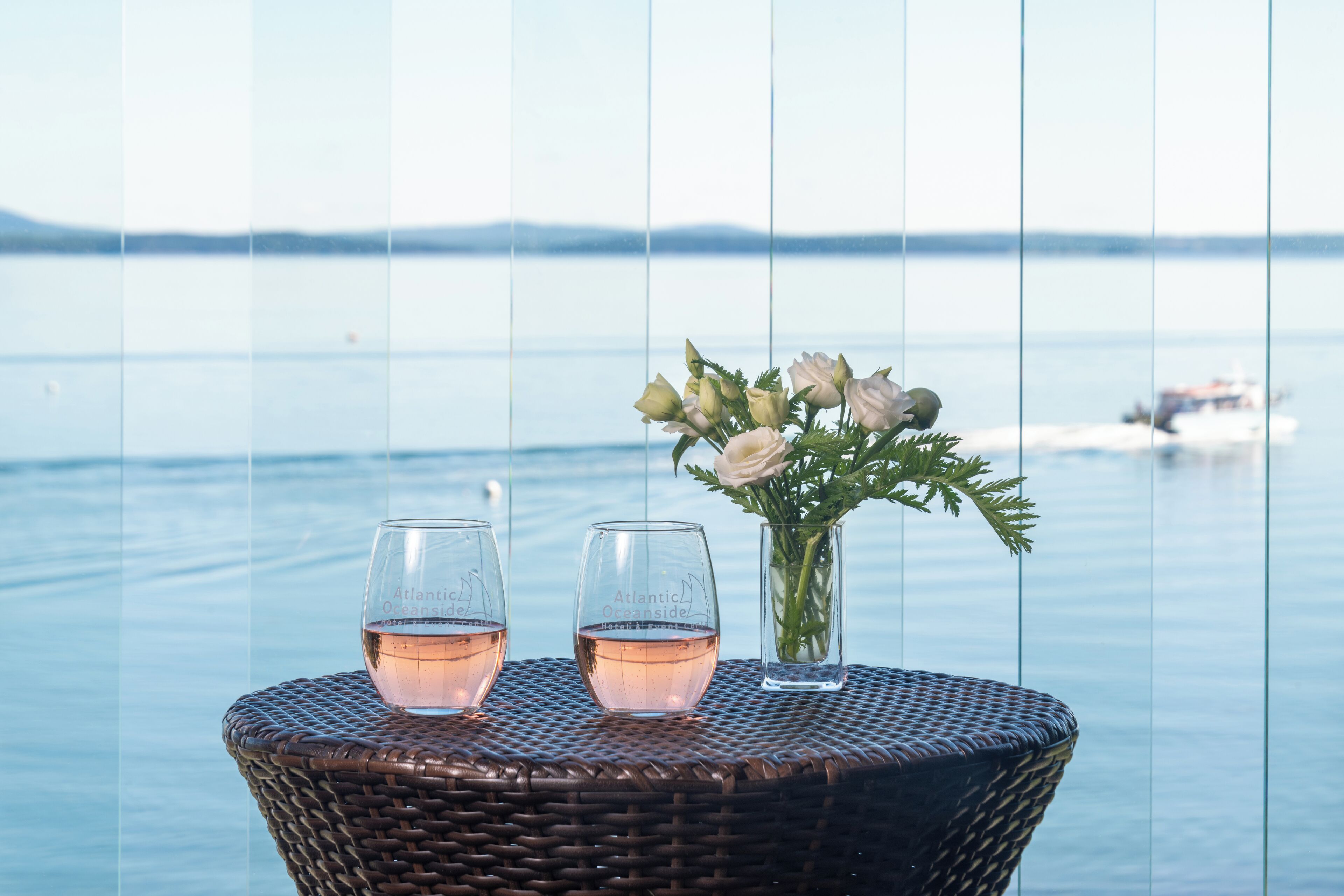 A wicker table with two glasses of pink wine and a vase with white flowers, overlooking a calm body of water with a boat in the distance.