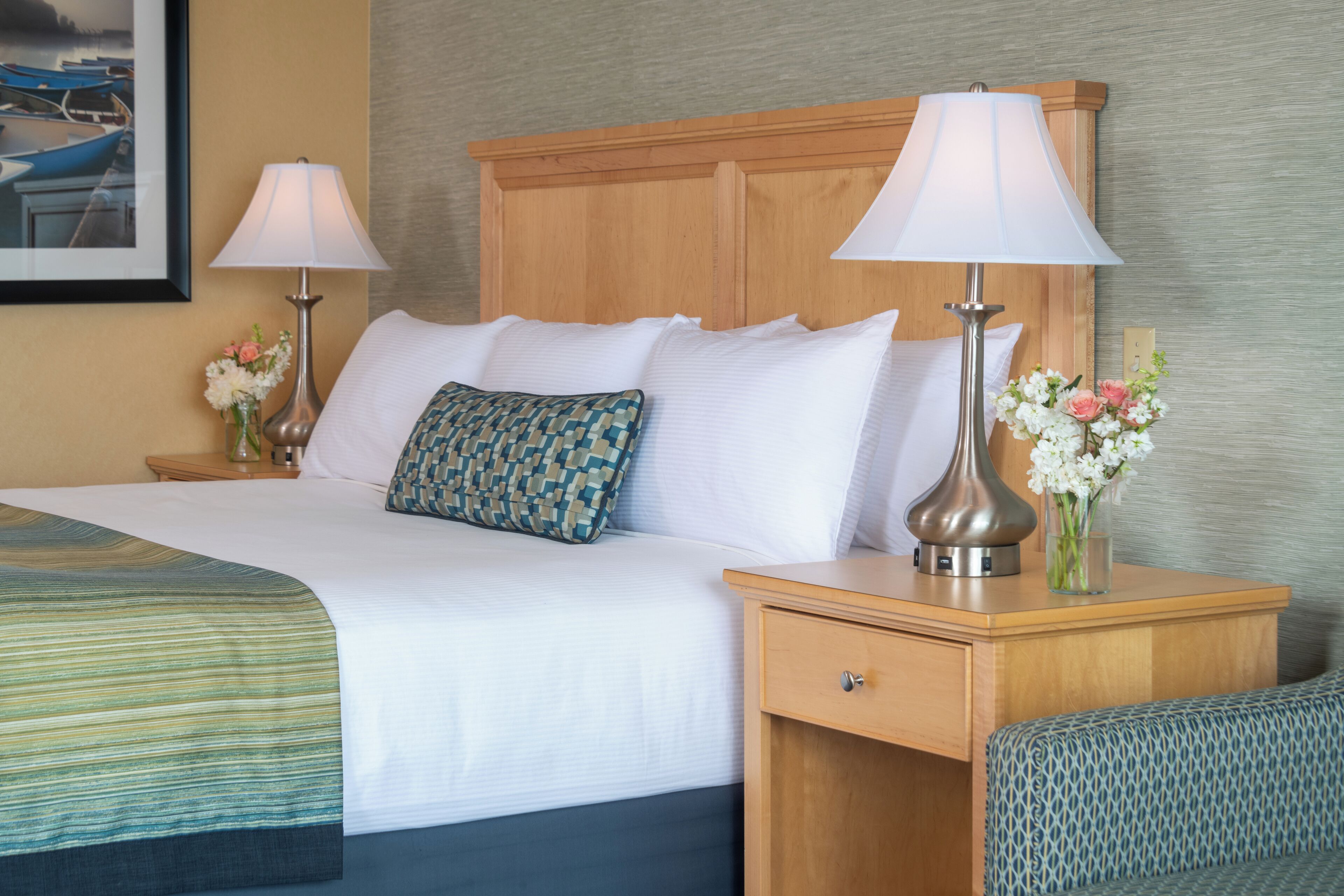 Cama bem arrumada com lençóis brancos e almofada estampada, com duas luminárias de cabeceira e um vaso de flores ao lado.