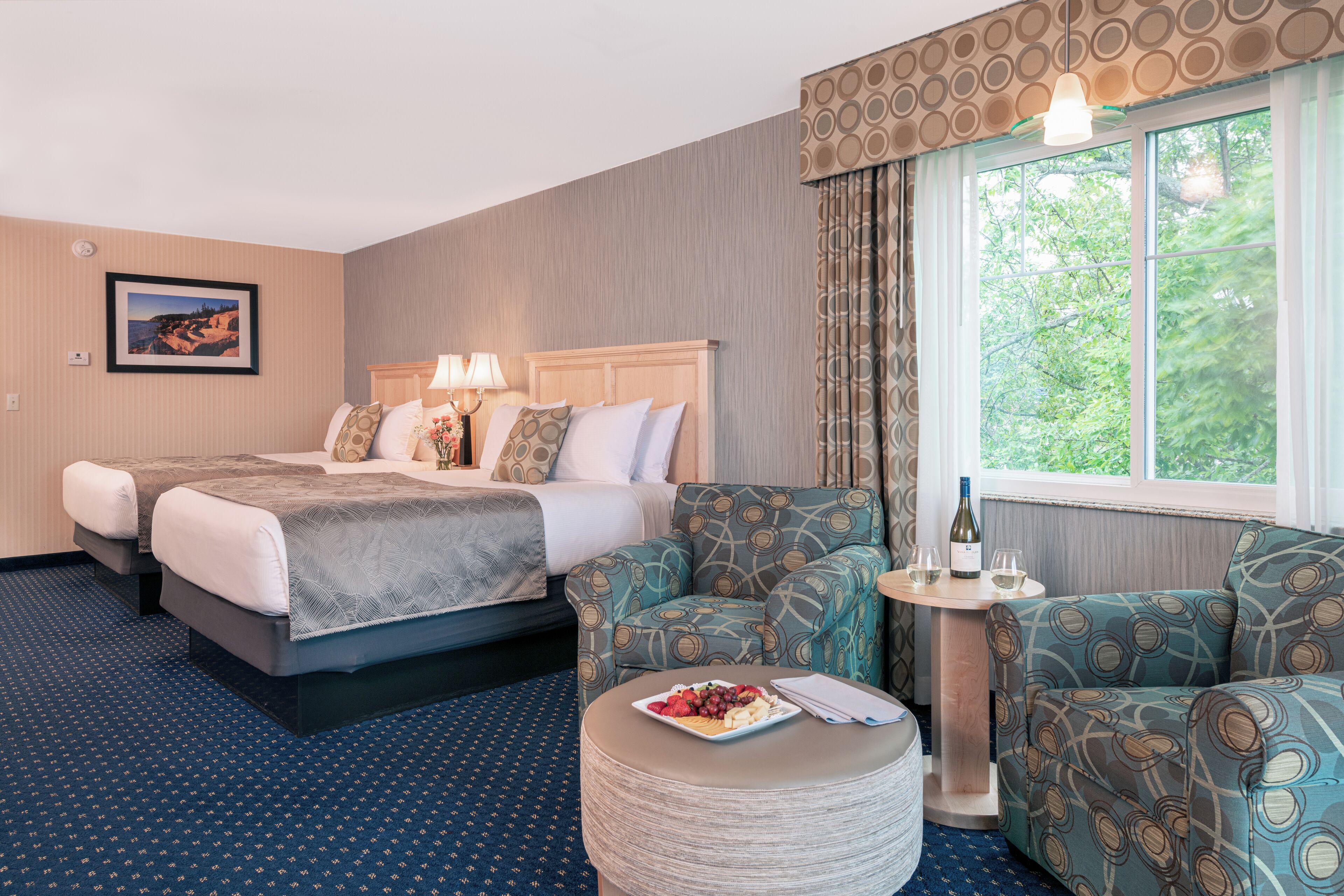 A hotel room with two beds, a round dining table, and a view of trees through the window.