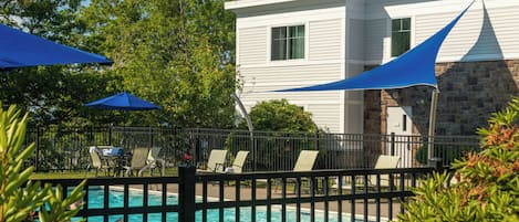 A swimming pool with lounge chairs and umbrellas, surrounded by a stone wall and a white building with windows.