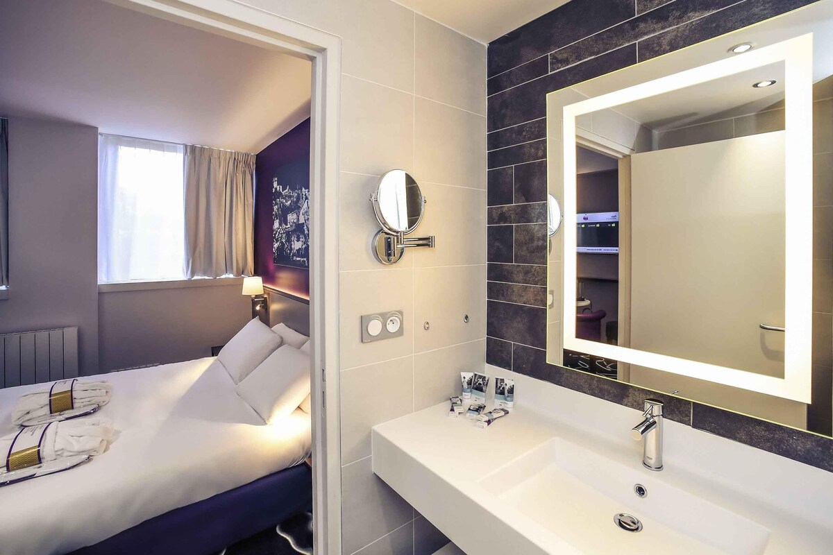 A modern hotel room with a bed, a large mirror, a bathroom with a sink and a shower, and a view of the city through the window.