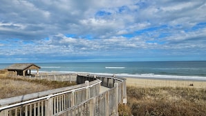 Une promenade en bois mène à une plage où se trouve une petite construction en bois, d’où l’on peut admirer l’océan sous un ciel partiellement nuageux.